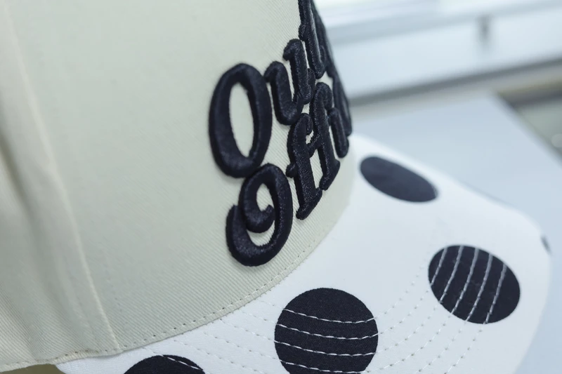 Close-up of black raised embroidery, visor stitching and black polka-dot pattern on a cream baseball cap